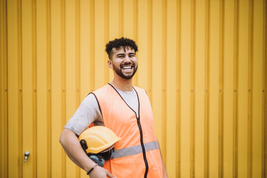 Portrait of cheerful young construction worker with hardhat standing against yellow metal wall