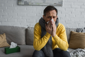 ill man in warm scarf sneezing in paper napkin with closed eyes while sitting on couch.