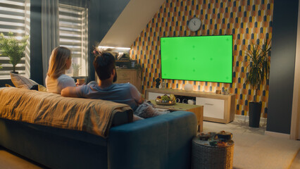 Couple sitting on sofa in living room, talking, watching breaking news, movie or series on TV. Wife and husband resting and chilling at home in the evening together. Green screen. Chromakey.