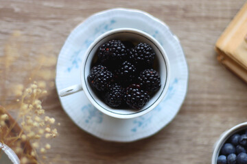 Vintage tea set filled with blueberries, blackberries and starwberries, open book, reading glasses and decorative flowers on the table. Flat lay.