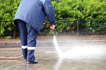 Worker in blue uniform watering the sidewalk with a hose. Street cleaning and disinfection in...