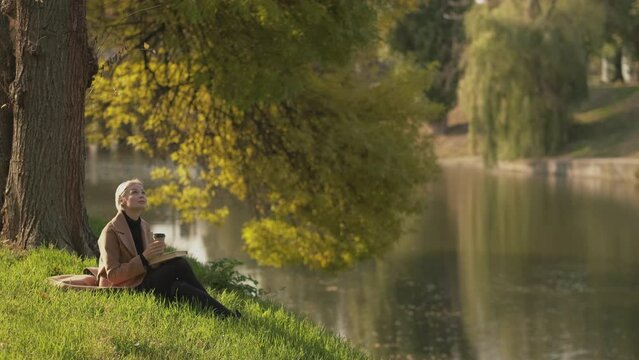 Autumn inspiration. Romantic woman. Pleasant leisure. Pretty happy middle-aged lady sitting lake shore enjoying beautiful nature with cup of coffee and book.