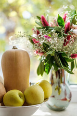 Close up shot of an autumn composition, bouquet of flowers in a vase, butternut squashes, apples in a bowl.