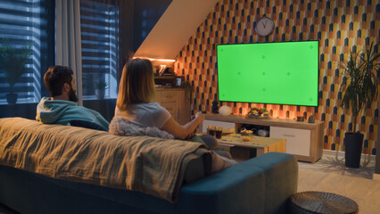 Woman switching channels with remote control. Couple sitting on couch in living room, talking, watching breaking news, movie or series on TV. Resting at home in the evening. Green screen. Chromakey.