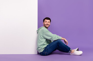 Full length photo of nice young man sit floor look interested white wall dressed stylish khaki...