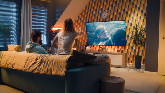 Happy Couple Sitting On Sofa, Watching Action Movie On TV Or Criminal Blockbuster On Streaming Service, Talking And Discussing Acting, Eating Snacks, Pizza, Chips. Spouses Resting At Home In Evening.