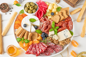 Charcuterie board with a variety of cheeses, salami, chorizzo, prosciutto, honey, grapes, nuts, olives, bread, blueberries and fresh herbs on a white wooden background. A festive snack.