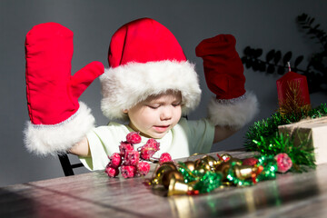 Fototapeta na wymiar A small child in a Santa Claus hat and red mittens with Christmas gifts is sitting at the New Year's table. Celebrating Christmas.