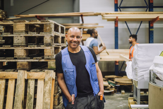 Smiling bald carpenter with hands in pockets leaning on wooden rack in warehouse