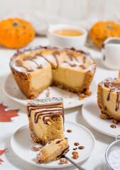 A piece of cheesecake on a white plate garnished with chocolate sauce. The spoon is broken off a small piece. Blurred in the background is a cheesecake and a pumpkin and a mug of tea. 