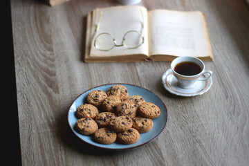 Plate of chocolate chip cookies, stack of vintage books, reading glasses, cup of tea or coffee, lit candle and fairy lights. Hygge at home. Selective focus.