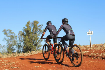 Ciclistas participando de competição em trilha de terra vermelha do brasil, em dia ensolarado com céu azul.