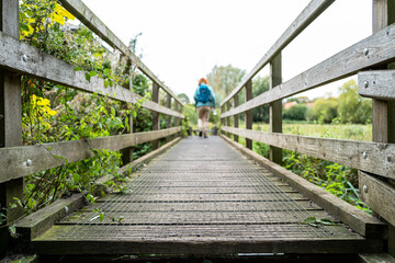 Surface level view of a distant trail walker seen crossing a slippery footbridge. The loan woman is seen with a day sack on her back.