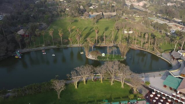 Drone video of a lake or lagoon. with a small island in the middle. Surrounded by grass and trees. Half of the lake is covered by shadow. Drone flies forward and tilts gimbal down.