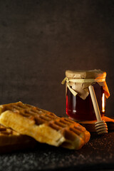 Photo still life with freshly baked waffles and a jar of honey with a honey stick on a dark...