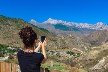 A young woman takes pictures on a phone from an observation deck of a rural settlement in a mountain gorge.