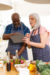 Happy senior diverse couple wearing aprons and using tablet in kitchen