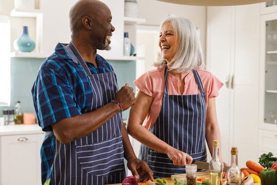 Happy senior diverse couple wearing aprons and cooking in kitchen - Powered by Adobe