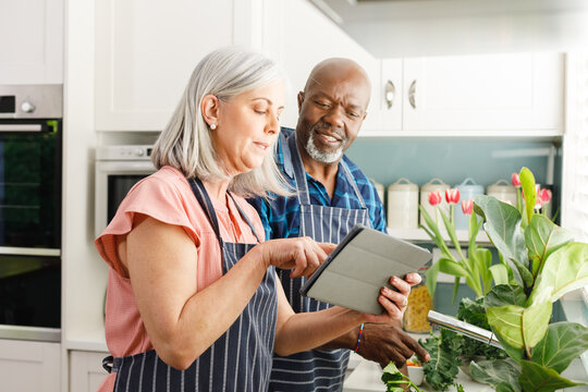 Happy senior diverse couple wearing aprons and using tablet in kitchen