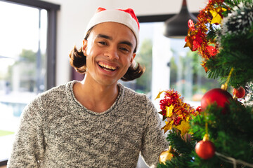 Happy biracial man wearing santa claus hat, decorating christmas tree in living room