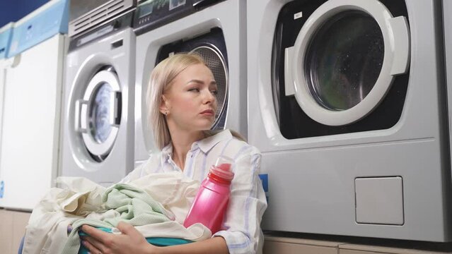 A woman is sitting near a household washing machine, which is located in a public laundry, she is waiting for clean laundry