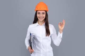 Smiling woman in protective helmet with clipboard isolated on grey background. Young woman construction manager. Architect woman with ok sign, female worker in hardhat helmet.