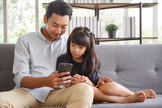 Cute asian girl help young father show something on smartphone making selfie together, smart daughter and dad sit on couch hold mobile phone take family picture on moblie phone.