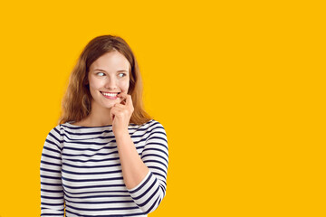 Young woman looks aside with funny awkward smile on face. Beautiful lady standing on yellow studio background looks at blank copy space side, sees something, thinks of guilty pleasure, feels nervous - obrazy, fototapety, plakaty