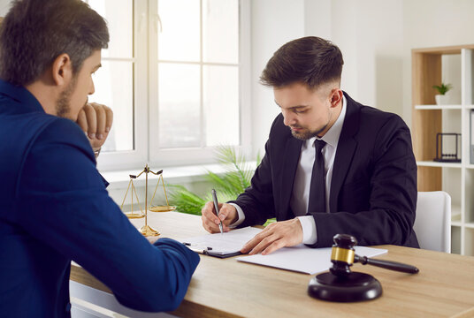 Lawyer working with a client. Professional lawyer, attorney or legal advisor sitting at his office table with a gavel and scales of justice and signing some documents. Law, legal services concept