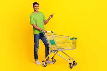 Full size photo of positive man market trolley indicate finger empty space isolated on yellow color...