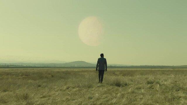 4K Back rear view of lonely man walking on field . Young stylish male artist standing on meadow field at day time . Concept of fantasy mystic dreams . Approach of two nearby planets . 