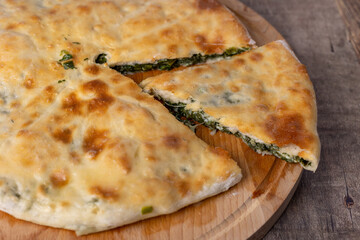Pie with spinach, herbs and suluguni cheese on a wooden board. Whole pie with cut off piece. Traditional round flat pie with filling. Homemade pastries of Caucasian and Balkarian cuisine. 
