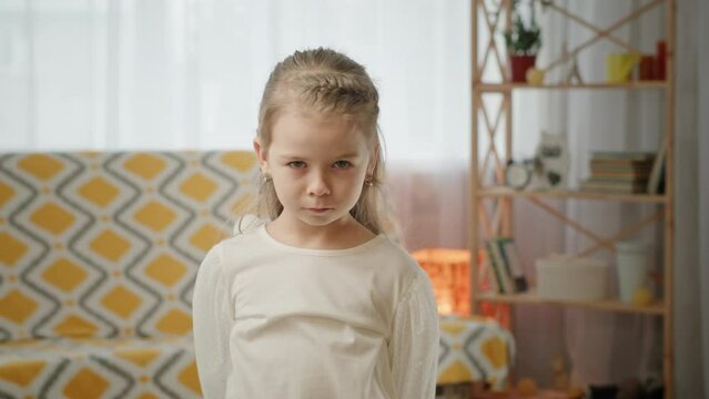 Preschooler girl stands with lowered head and looks offended. Gloomy preschooler girl in white dress stands with lowered head in light room. Blonde child looks forward and imitates offended narrowing