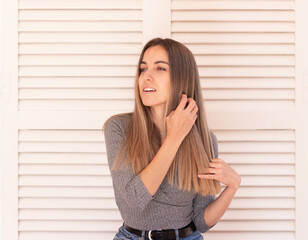 Young beautiful blonde woman touching and brushing her hair by hand while looking to the side in white door background