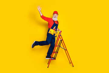 Fototapeta na wymiar Full length photo of funny positive old guy dressed uniform overall red hardhat rising stairs waving arm isolated yellow color background