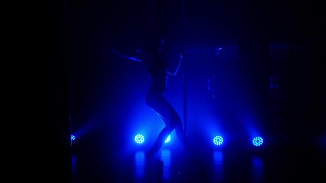 Slim woman performing poledance tricks sensually in spotlights. Sexy lady making moves upside down in night club. Young blonde dancer doing twine on pylon illuminated soft nightclub backlight.