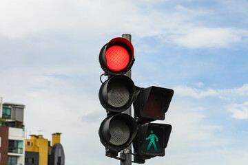 Vehicle traffic light in red, next to pedestrian which is green on a background between clouds and a piece of blue sky.