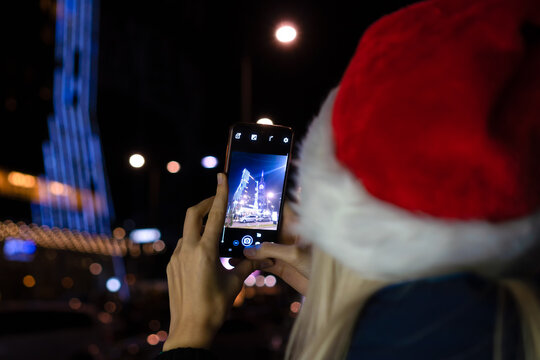 Young blonde woman in a Santa Claus hat takes pictures of the night city on a mobile phone, photo from the back