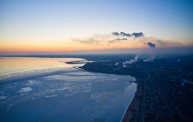 Aerial of industrial plant on the coast of a frozen Sea of Azov in Mariupol. Top view of the factory, smoke rising from the chimneys. City near icy ocean on winter sunset. Global warming concept.