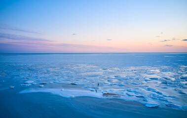 Naklejka na ściany i meble Aerial view - two people walk on ice on sunset over the frozen sea. Winter landscape on seashore during dusk. View from above of melting ice in ocean on sunrise. Global warming. Vivid colorful skyline