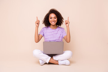 Portrait of positive gorgeous girl sit with netbook wear striped t-shirt indicating empty space isolated on beige color background