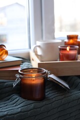 Burning candle in jar on knitted plaid near window indoors, space for text