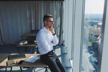 Cute brutal young businessman in glasses and white shirt, he is in modern office with big windows. A man in classic trousers and a white shirt. Young attractive businessman