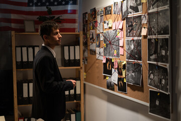 Detective looking on investigation board with photos of suspected criminals, crime scenes and...