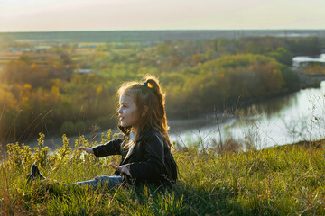 a little girl with curly hair sits on the ground on a walk outside the city in the rays of the setting sun