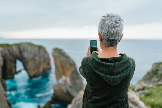 Senior woman with gray hair using her smartphone to take a landscape photo while hiking along a coastal path.