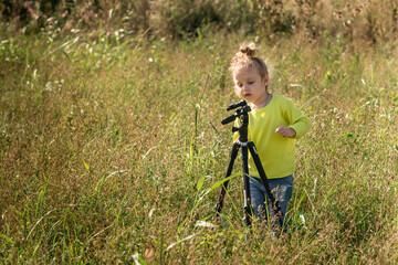 little inquisitive girl with blond wavy hair plays with a tripod for photos in the park in the warm...