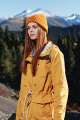 Woman in yellow raincoat hiking stands in front of mountains in yellow cap in autumn