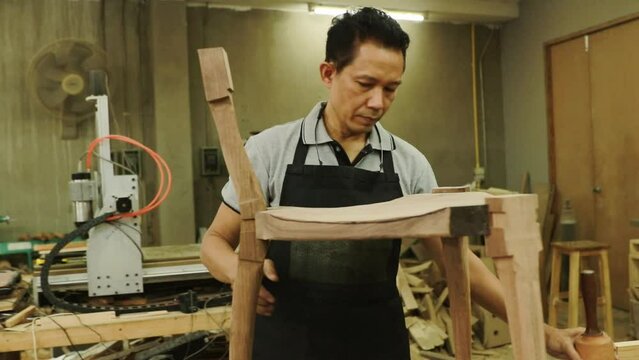 Senior asian carpenter works with wooden hammers to meticulously assemble wooden chairs and furniture in a wood factory.