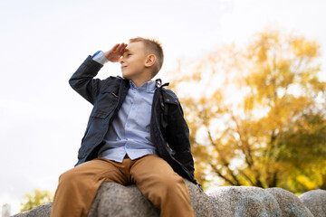 Fototapeta premium Cheerful mischievous boy sits on a stone and looks into the distance with his hand to his forehead